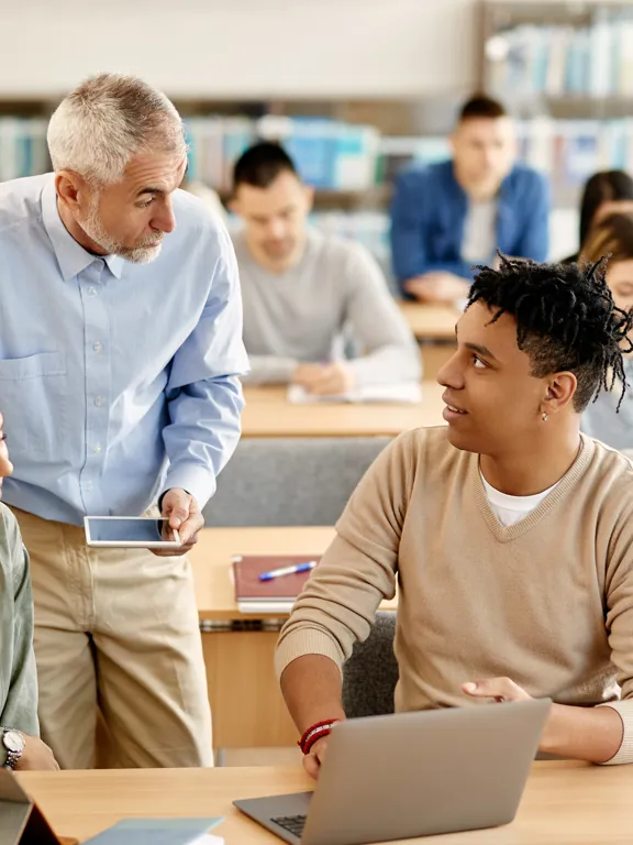 A male instructor with gray hair and a light blue shirt stands between two students sitting at desks in a crowded classroom or library, holding a small tablet and talking to a male student in a tan sweater. A female student in a green shirt is smiling on the left. Several other students are working in the background with shelves of books behind them.