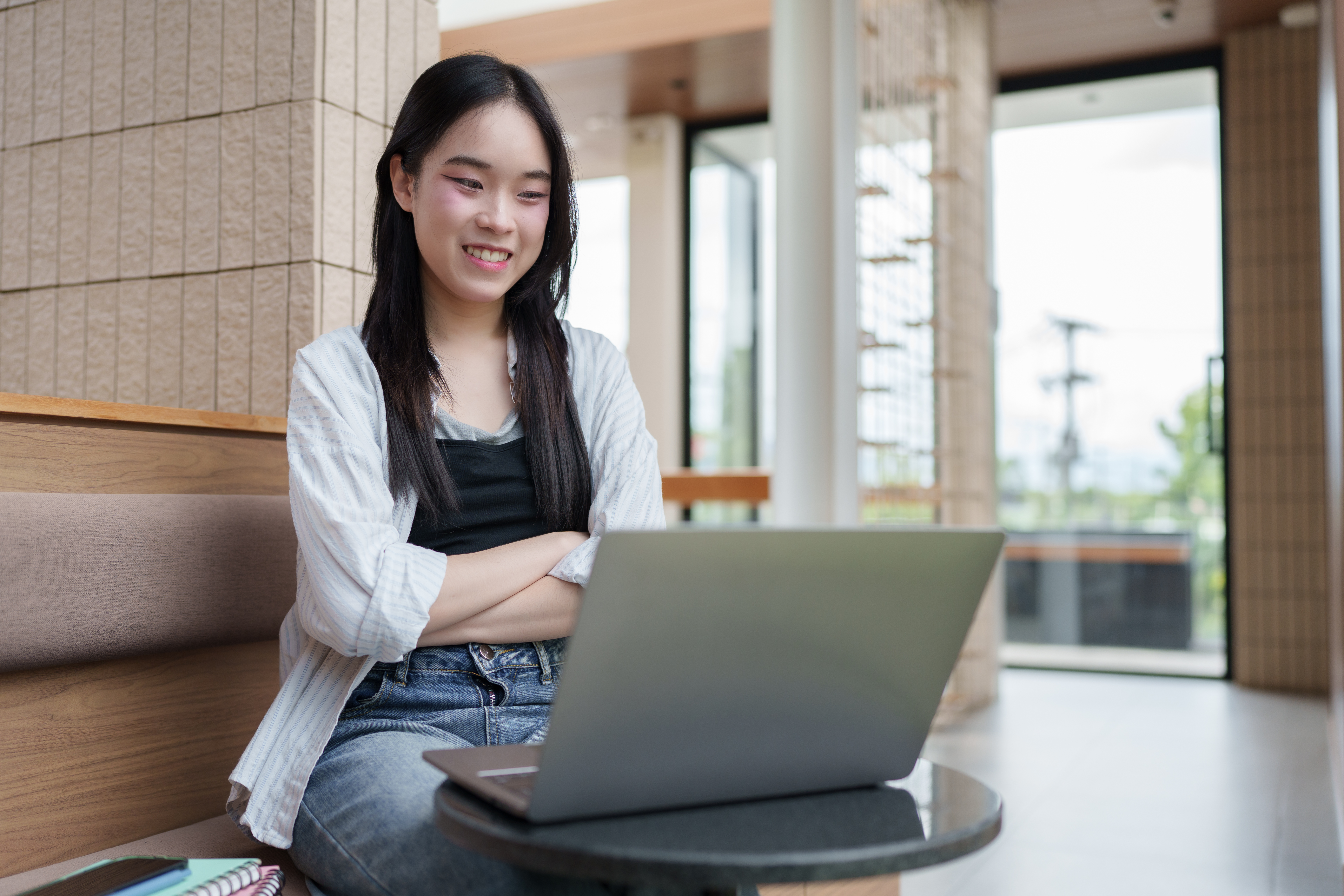 A smiling young woman with long dark hair sits comfortably with her arms crossed, looking at a laptop on a small table. She is wearing a white shirt over a black top and jeans, studying or working remotely in a modern, brightly lit indoor/outdoor common area.