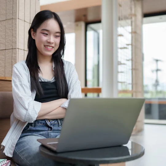 A smiling young woman with long dark hair sits comfortably with her arms crossed, looking at a laptop on a small table. She is wearing a white shirt over a black top and jeans, studying or working remotely in a modern, brightly lit indoor/outdoor common area.
