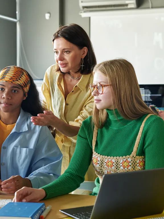 a female teacher assisting two female students working together on a computer in a modern classroom or computer lab.