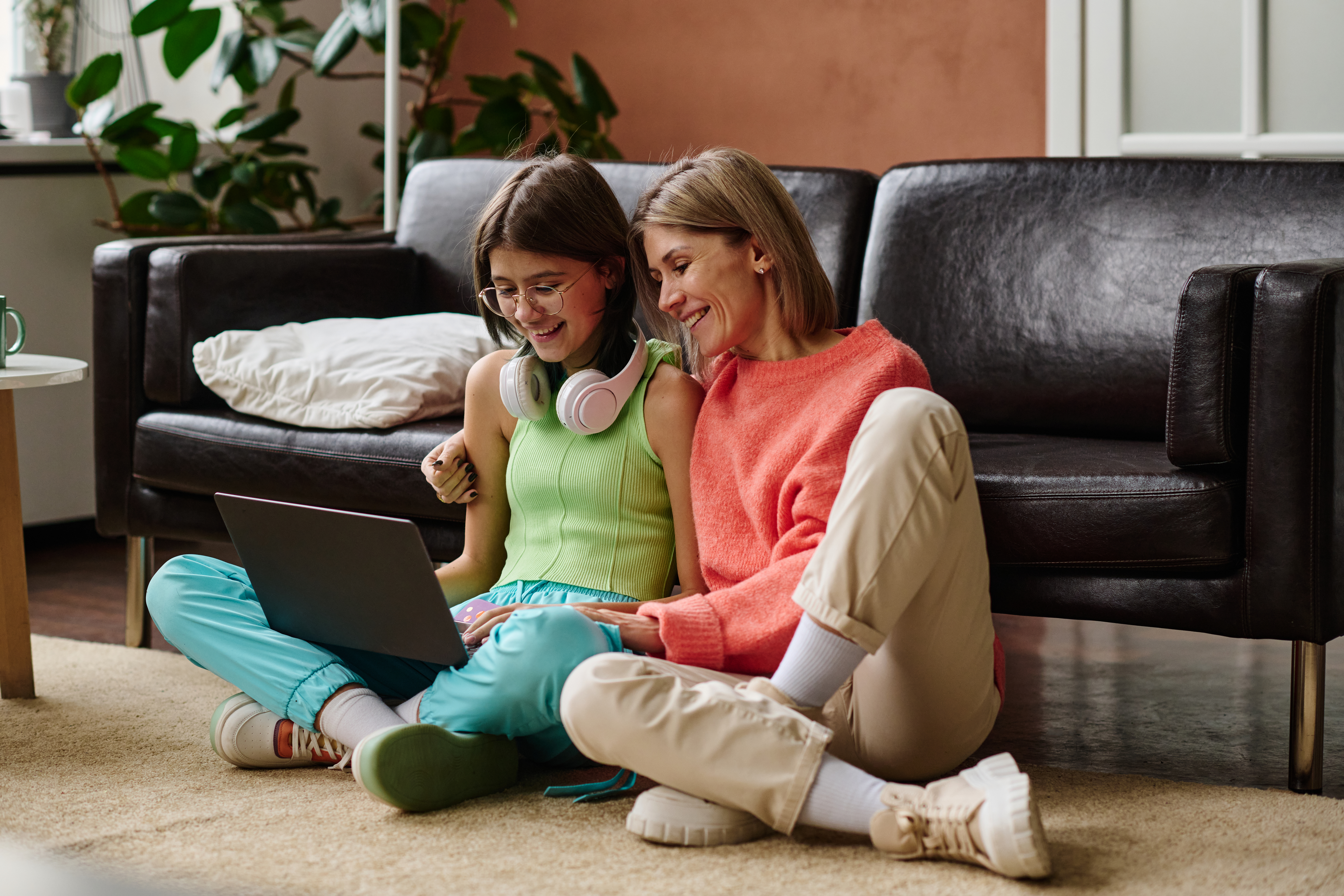 a smiling mother and daughter sitting on the floor, using a laptop together to look at the screen in a relaxed home setting.