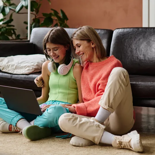 a smiling mother and daughter sitting on the floor, using a laptop together to look at the screen in a relaxed home setting.