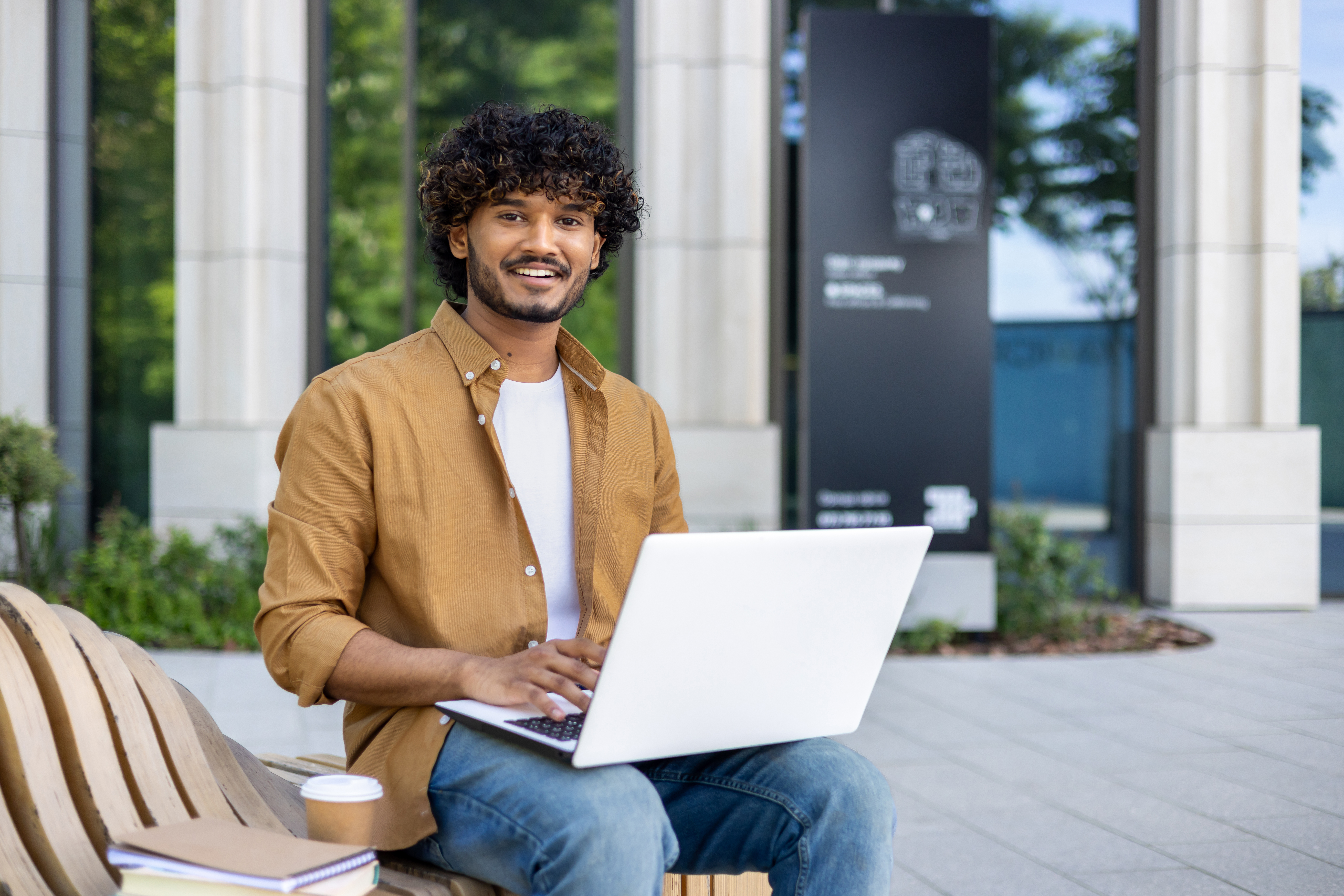 a young man smiling while working on a white laptop and sitting on a wooden bench outside a modern building.