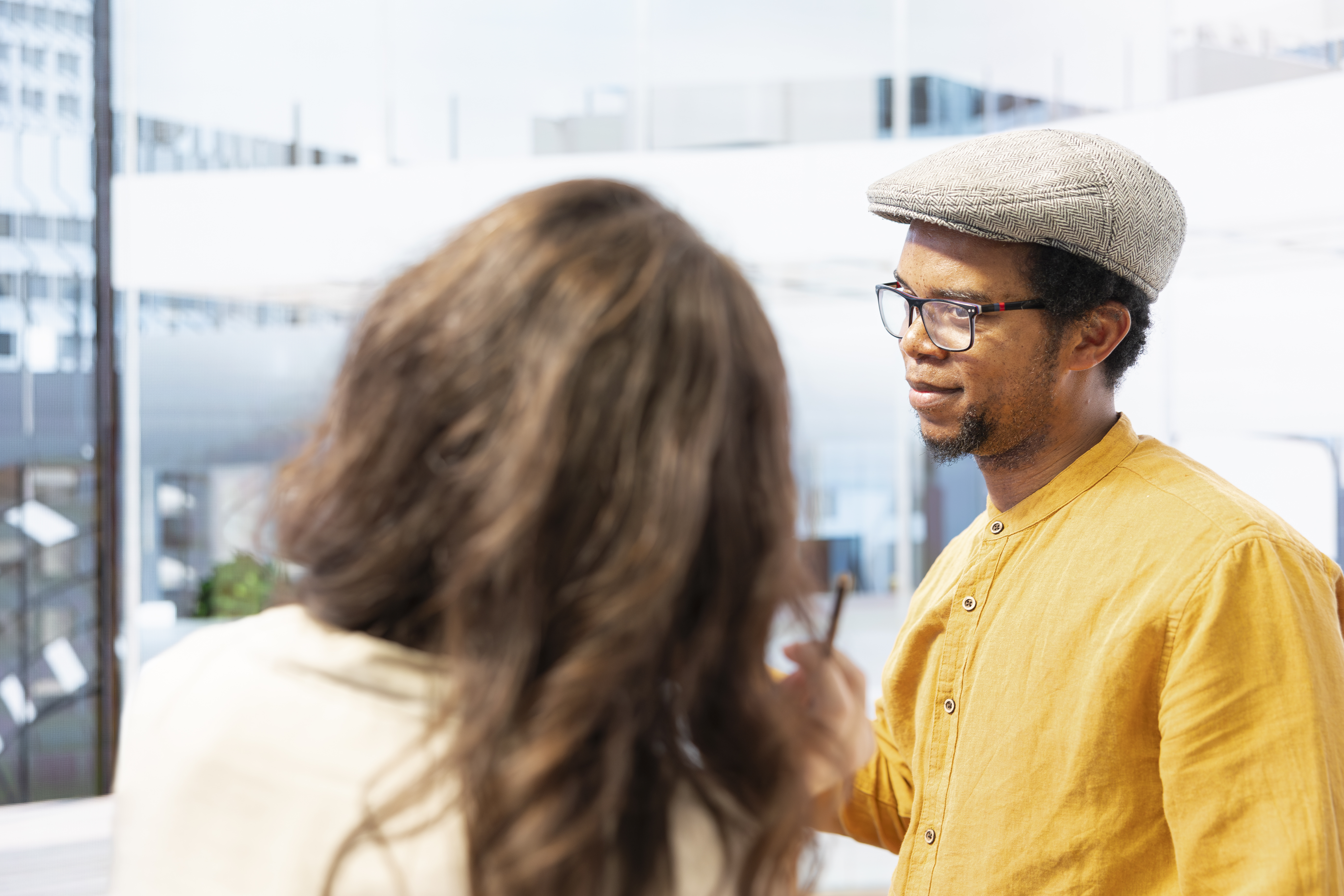 A man wearing a yellow shirt, glasses, and a flat cap is talking seriously with an unidentifiable person in a bright, modern office or study space.