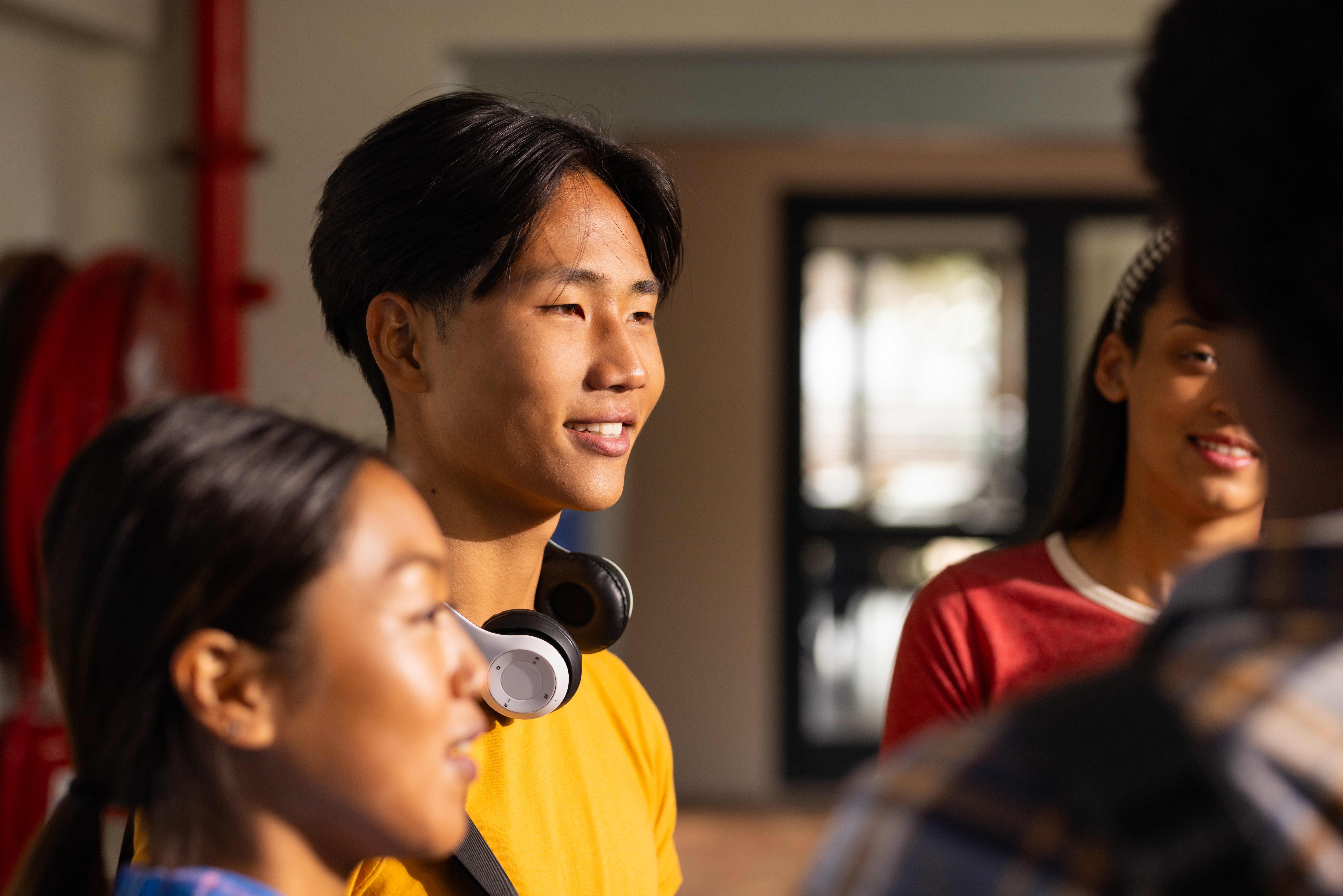 A smiling male student in a yellow shirt and headphones around his neck, engaged in a group conversation with other students on a school campus.