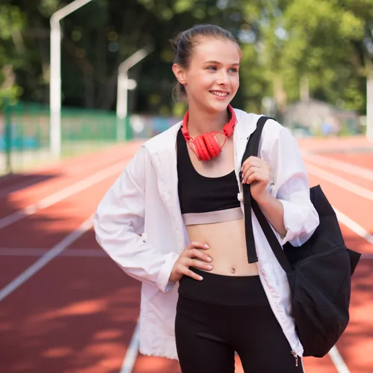 Smiling elite student-athlete standing on a running track with a sports bag and headphones. This represents the flexibility of online learning that empowers elite performers and athletes to manage rigorous training and competition schedules while achieving academic success.