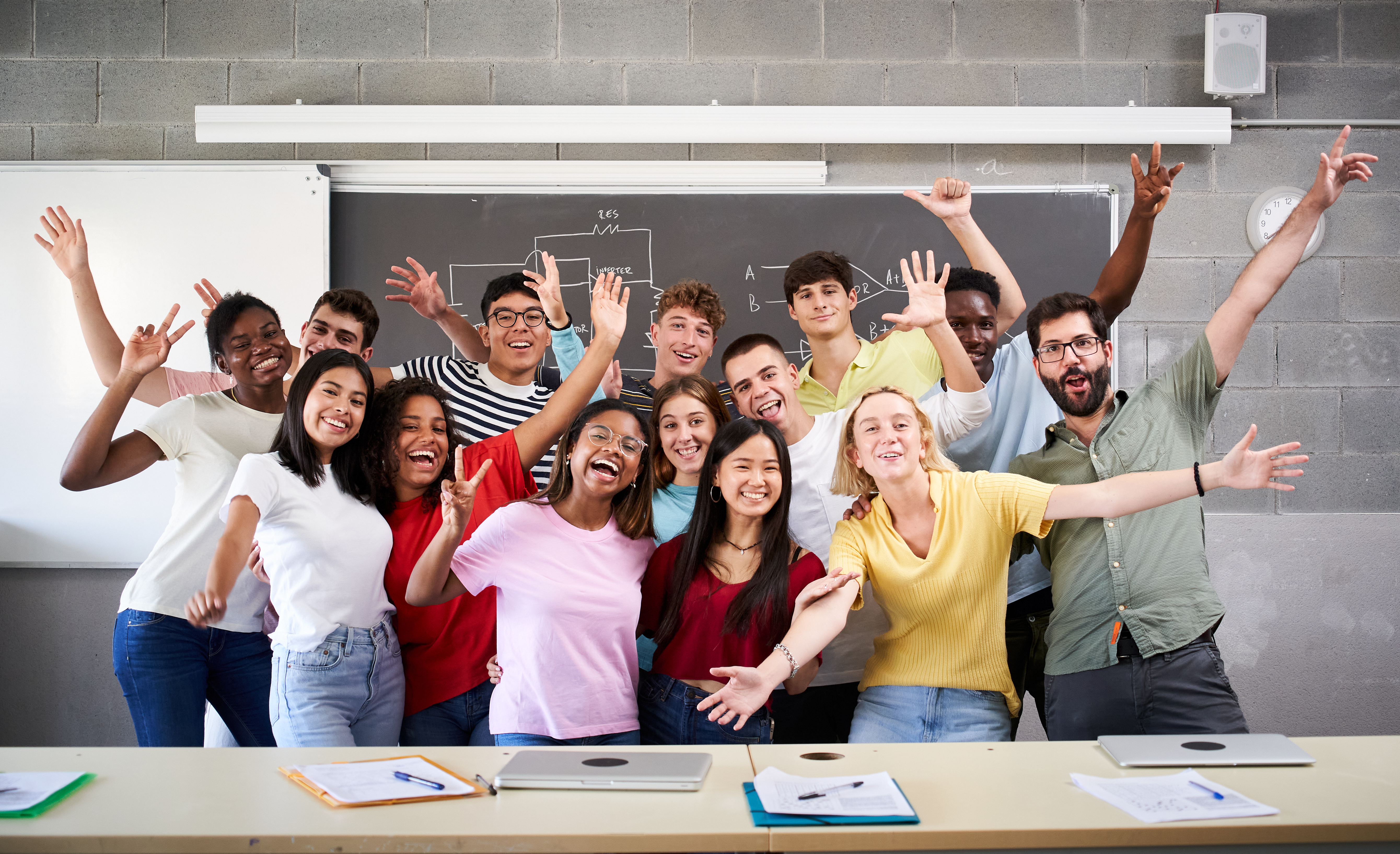 A large, diverse group of students and a teacher cheering and raising their hands in a classroom, with a diagram on the blackboard behind them.