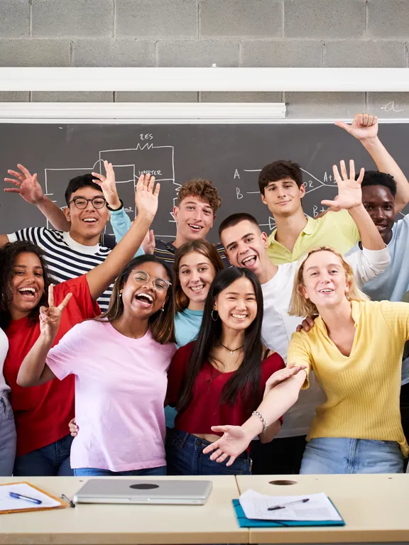A large, diverse group of students and a teacher cheering and raising their hands in a classroom, with a diagram on the blackboard behind them.