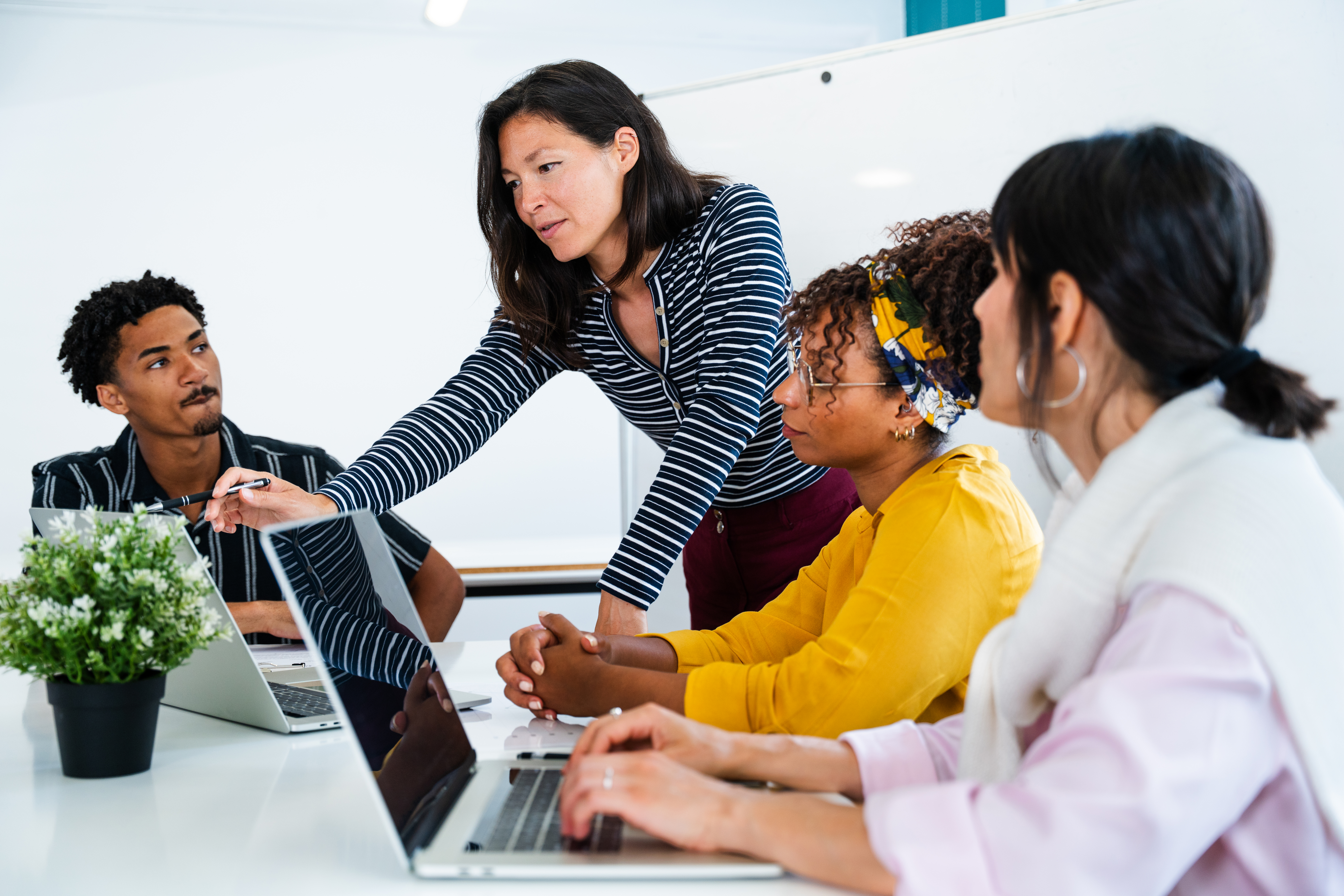 A female instructor in a striped shirt leans over a table, helping a diverse group of three female and male students working on laptops in a modern, bright setting.