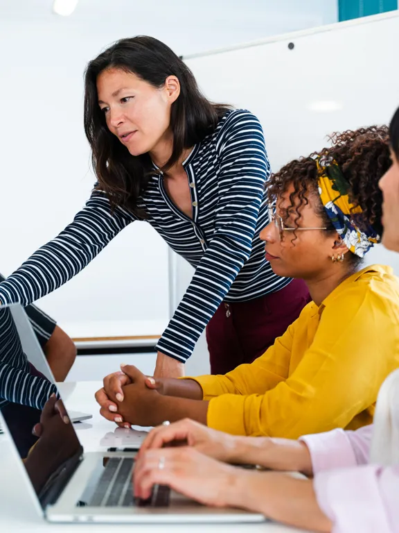 A female instructor in a striped shirt leans over a table, helping a diverse group of three female and male students working on laptops in a modern, bright setting.