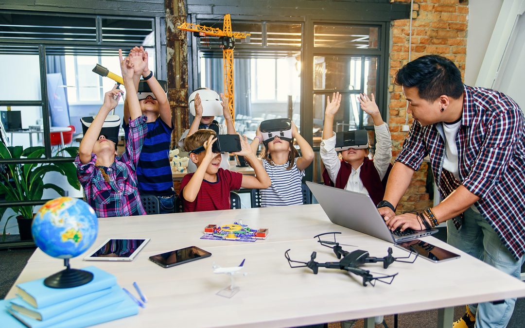 A teacher is standing with students who are seated at a table. The teacher is looking at a laptop. The students are wearing virtual reality headsets and have their hands raised in the air. On the table, there is a globe, notebooks, tablets, phones, a drone, and a toy airplane.