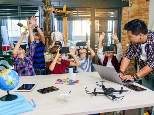 A teacher is standing with students who are seated at a table. The teacher is looking at a laptop. The students are wearing virtual reality headsets and have their hands raised in the air. On the table, there is a globe, notebooks, tablets, phones, a drone, and a toy airplane.