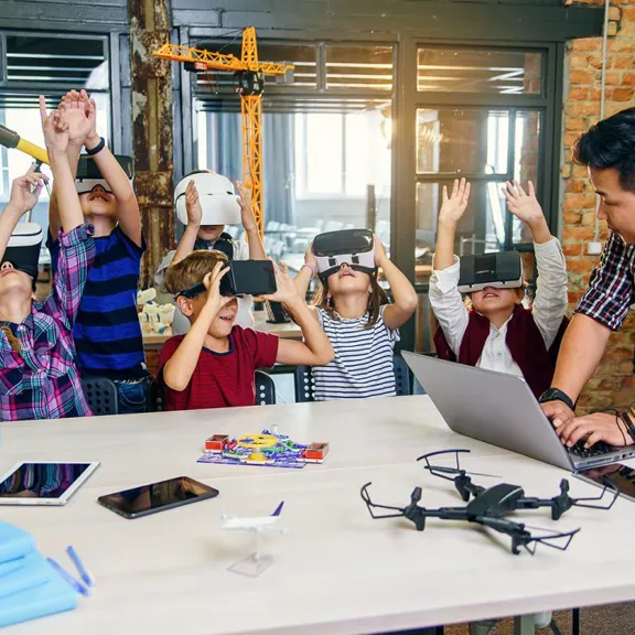 A teacher is standing with students who are seated at a table. The teacher is looking at a laptop. The students are wearing virtual reality headsets and have their hands raised in the air. On the table, there is a globe, notebooks, tablets, phones, a drone, and a toy airplane.