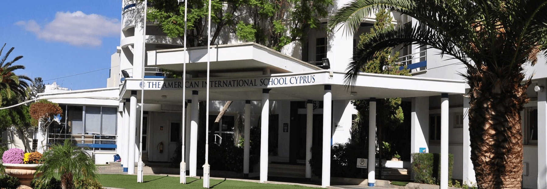 The main entrance to The American International School Cyprus, featuring a white covered portico with columns, and surrounding palm trees and greenery.