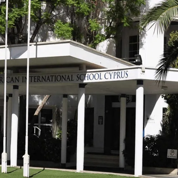 The main entrance to The American International School Cyprus, featuring a white covered portico with columns, and surrounding palm trees and greenery.