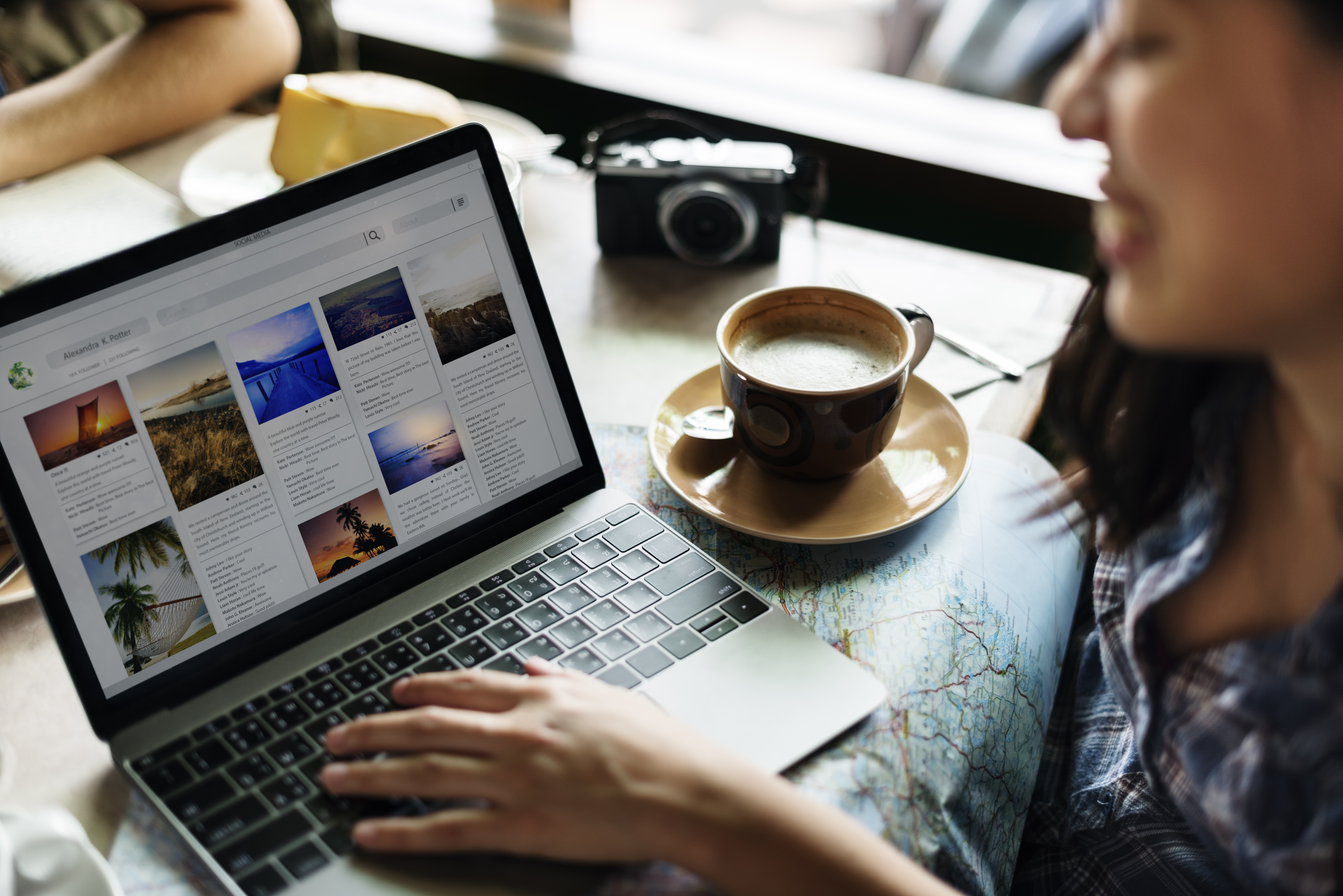 A person working on a laptop, displaying a travel blog or photo grid, with a cup of coffee and a map on the table in a café.