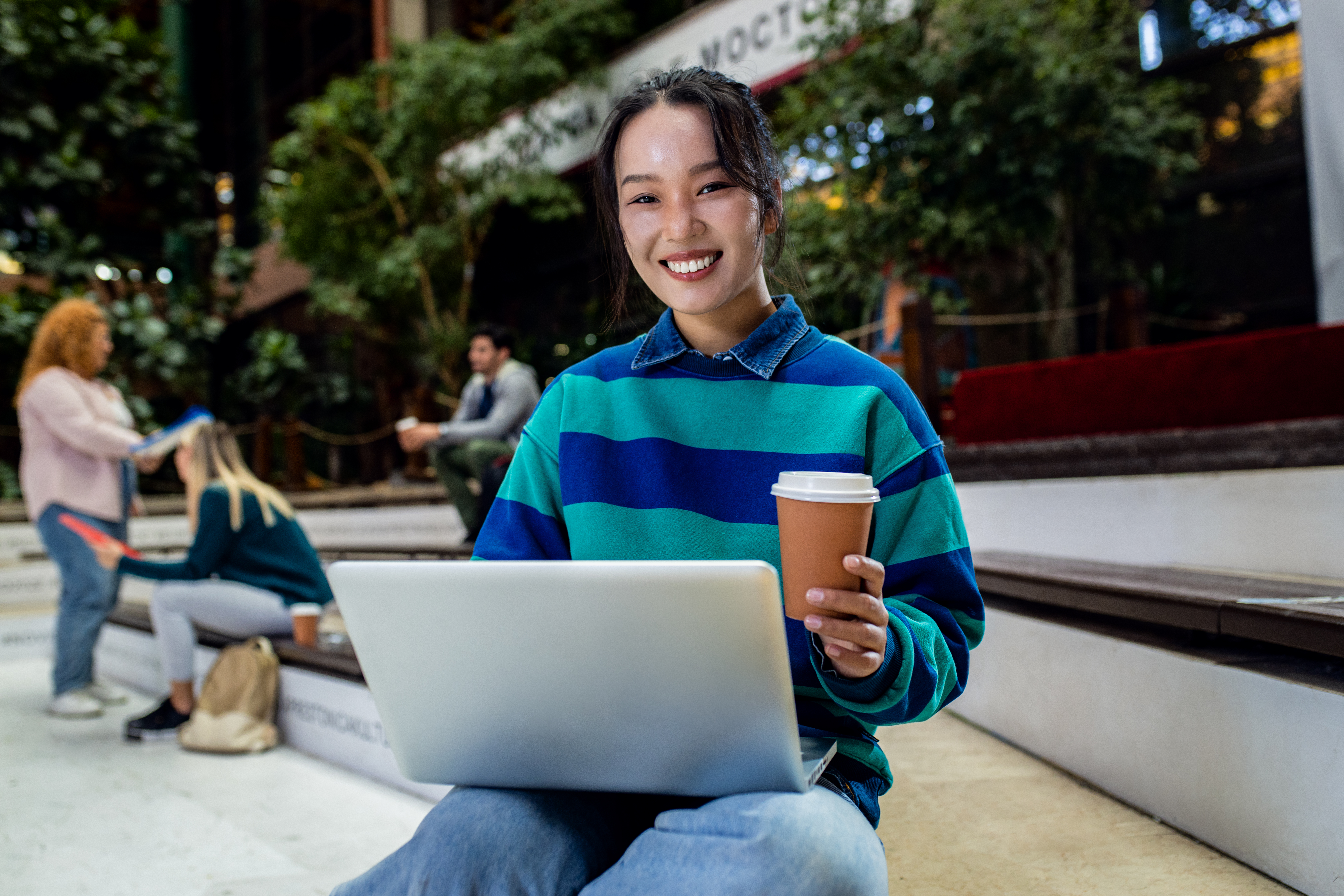 A young woman in a striped sweatshirt smiles while working on a laptop and holding a coffee cup in an outdoor public space, representing the mobility of digital society online learning.