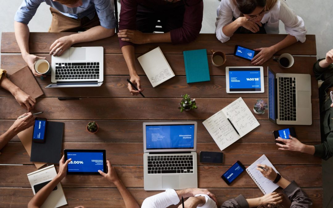 An overhead shot of a group of people sitting around a large wooden table. They are using various devices, including laptops, tablets, and smartphones, and have notebooks and coffee mugs on the table.