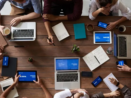 An overhead shot of a group of people sitting around a large wooden table. They are using various devices, including laptops, tablets, and smartphones, and have notebooks and coffee mugs on the table.