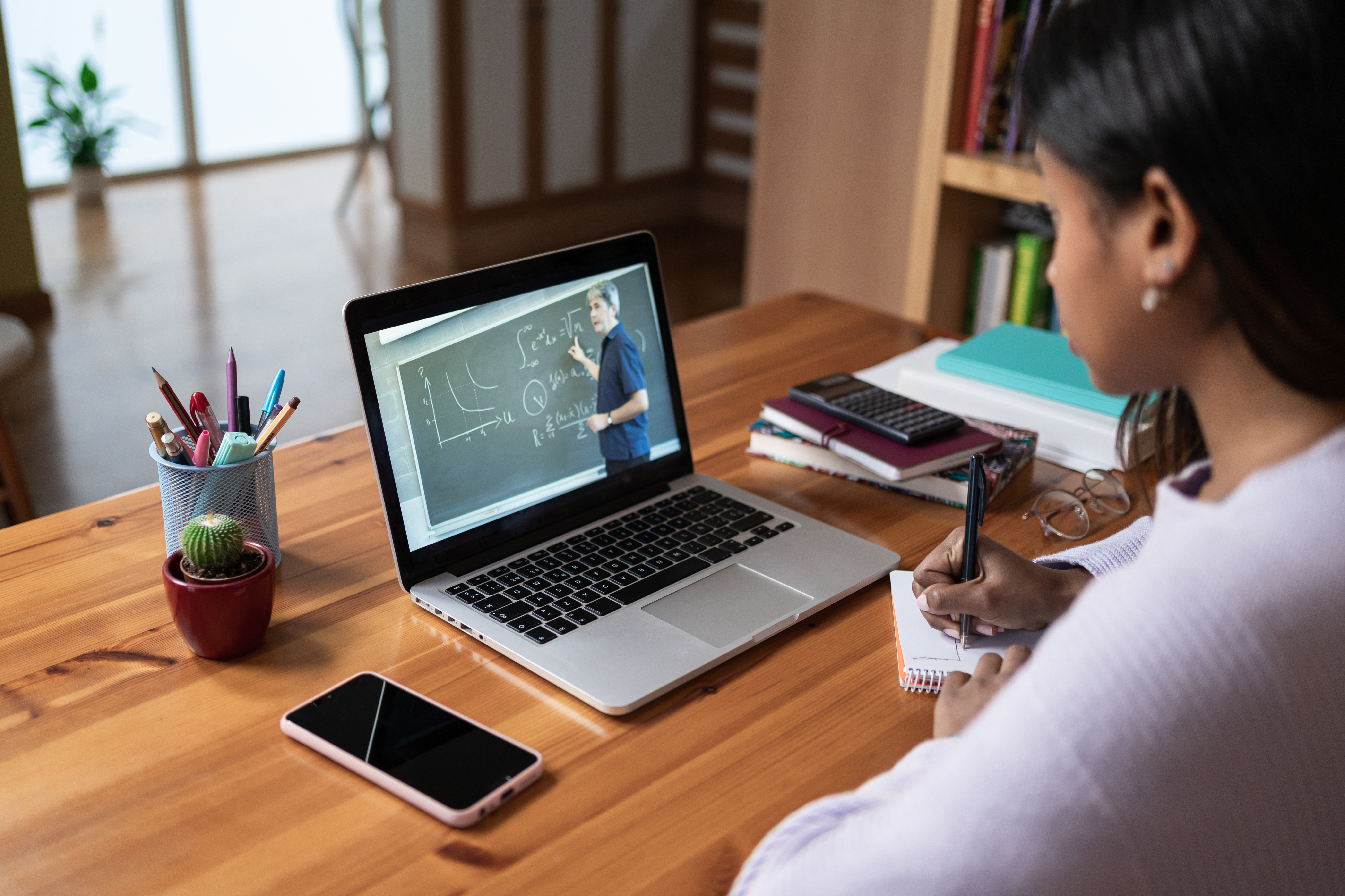 A student in front of a laptop, watching a teacher deliver a lesson in front of a classroom chalkboard.
