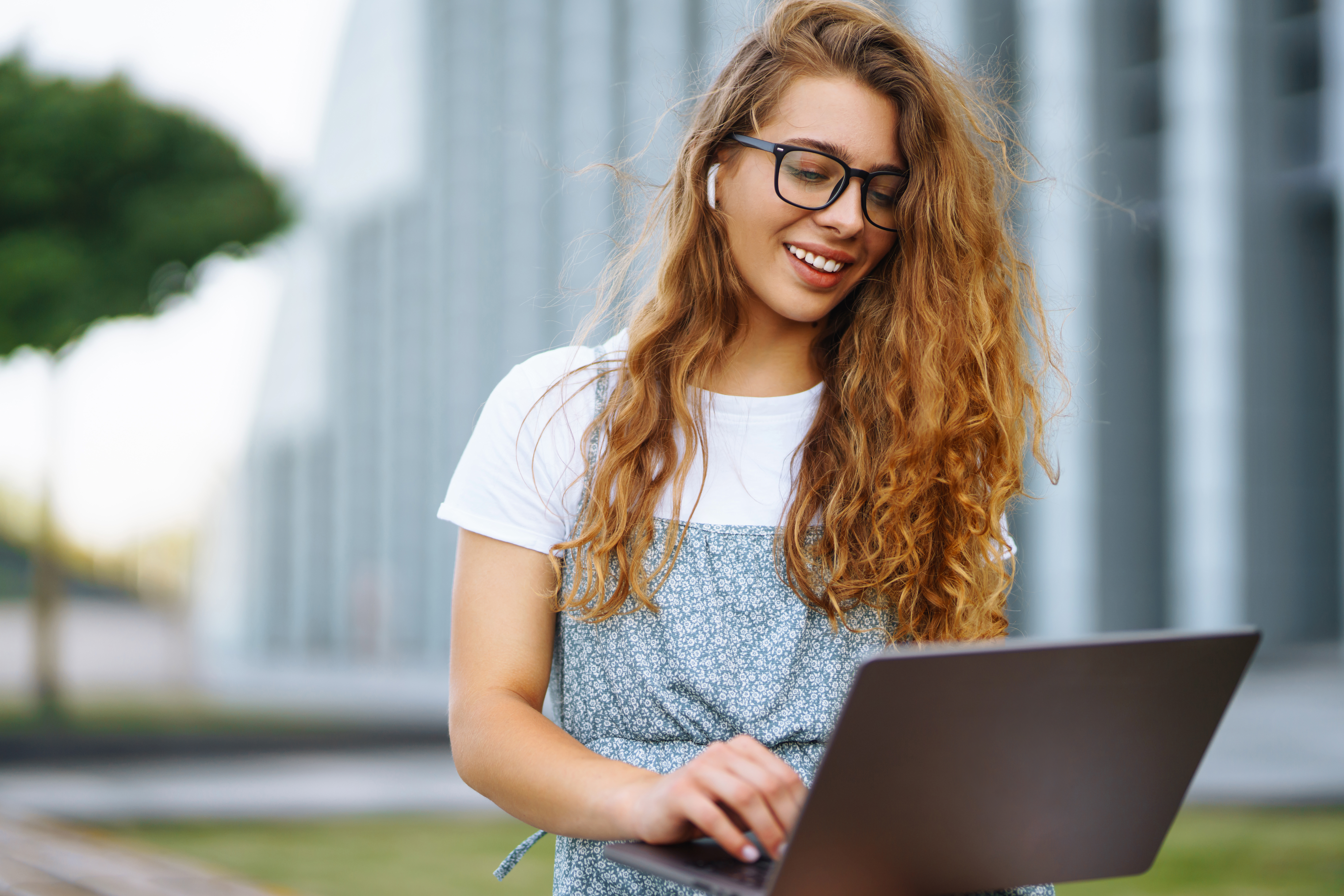 A happy young woman with long, curly auburn hair, wearing glasses and a patterned dress, works on a laptop outdoors. She has wireless earbuds in her ears and is smiling while looking down at the screen, with a modern building in the blurred background.