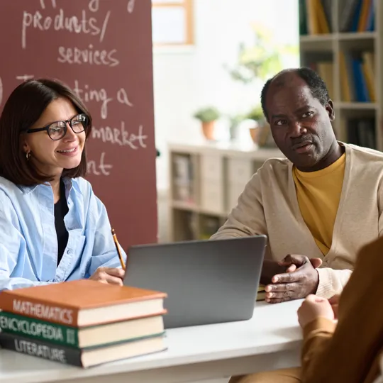 Three people, two adults and one seen from behind, discuss content on a laptop at a table with books and a chalkboard in the background.