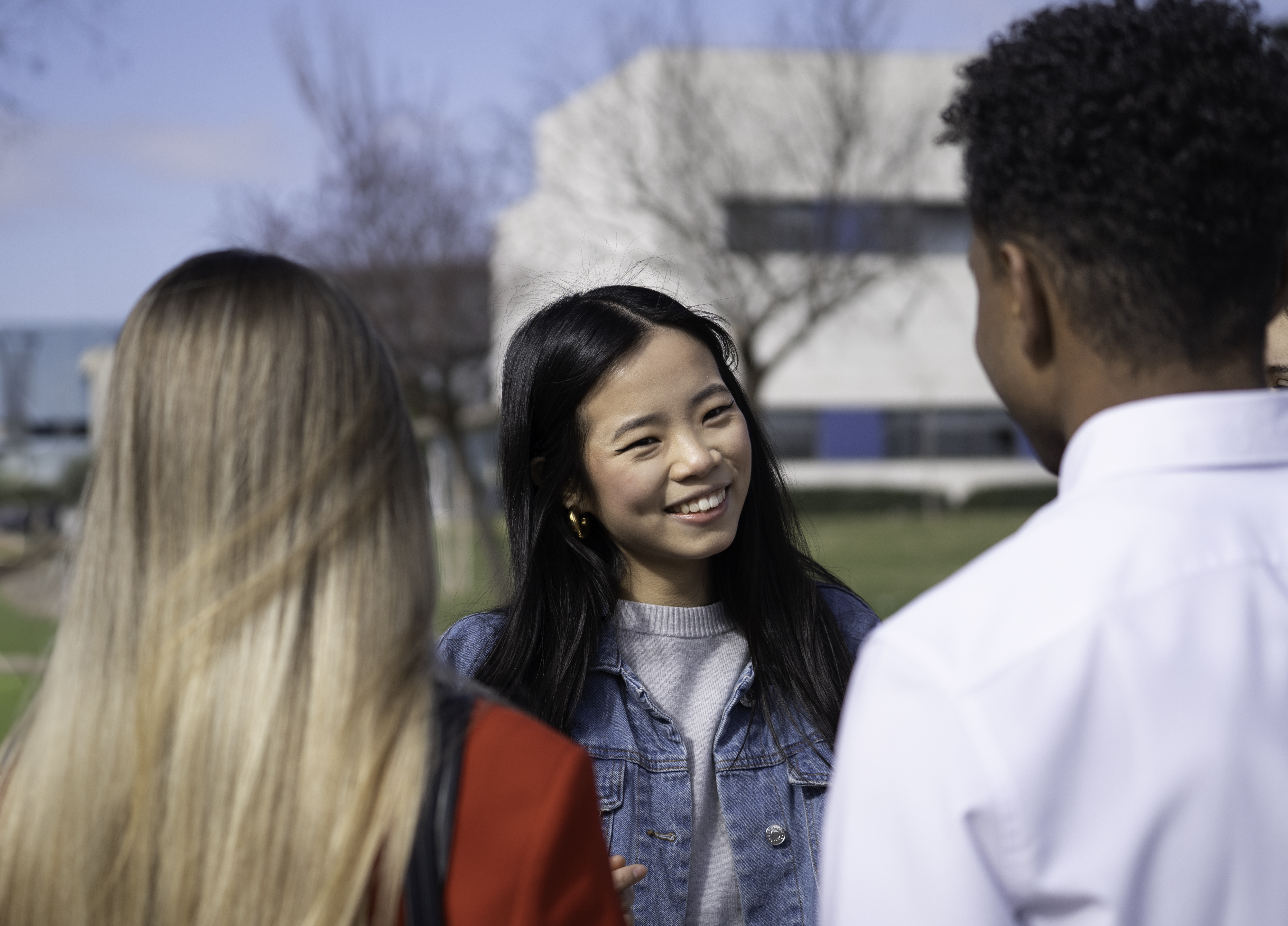 A young woman with long dark hair and a denim jacket smiles while talking to two classmates outdoors on a sunny university campus.