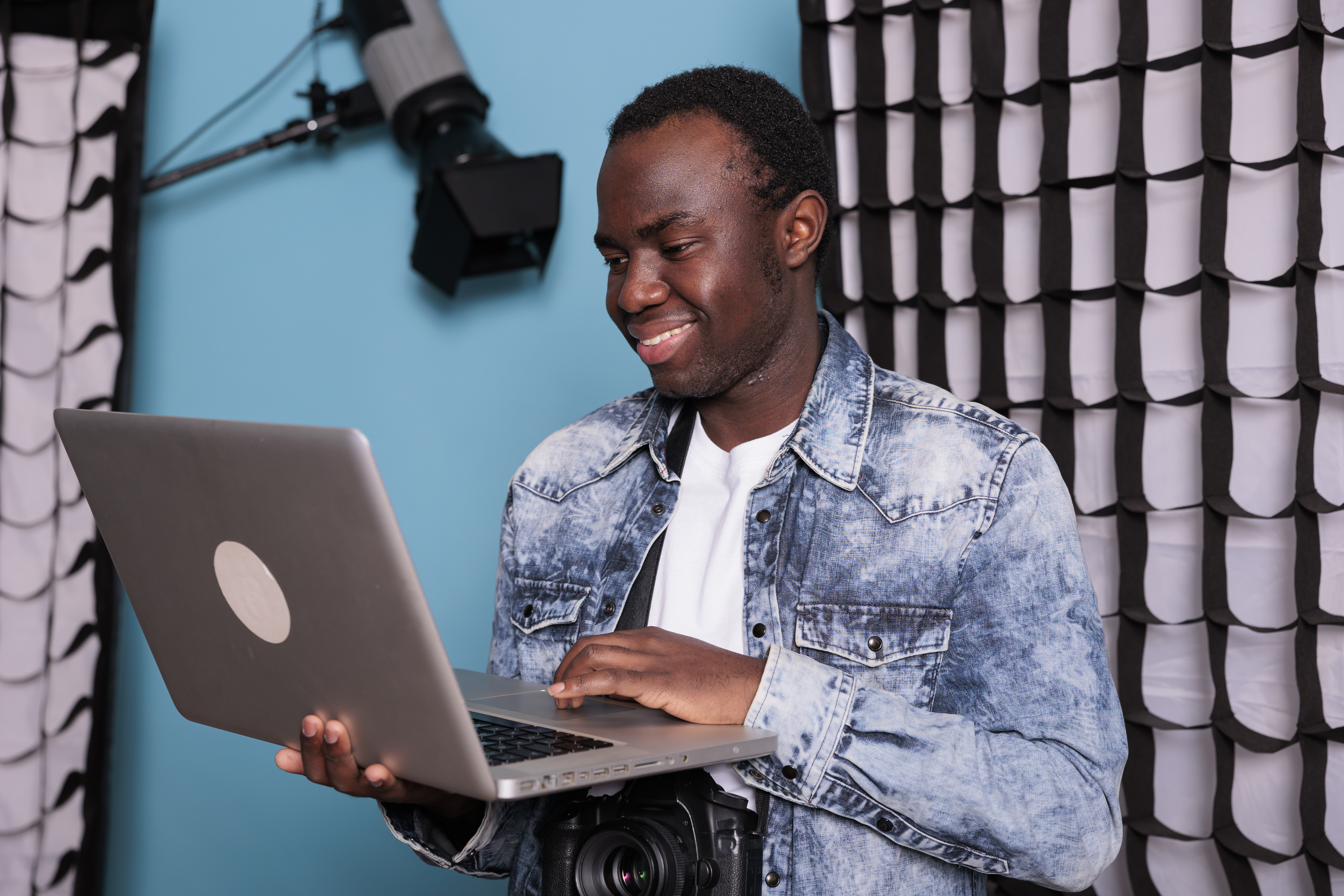 A smiling young man in a denim jacket works on a laptop while standing in a photography studio with a blue backdrop, representing flexible online learning.