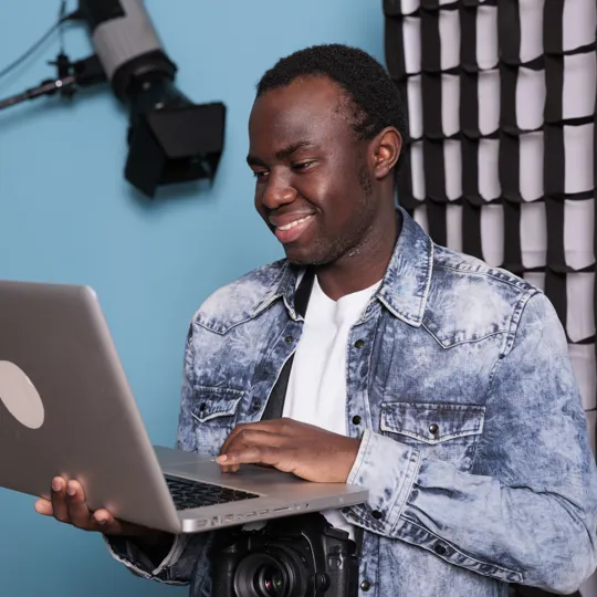 A smiling young man in a denim jacket works on a laptop while standing in a photography studio with a blue backdrop, representing flexible online learning.