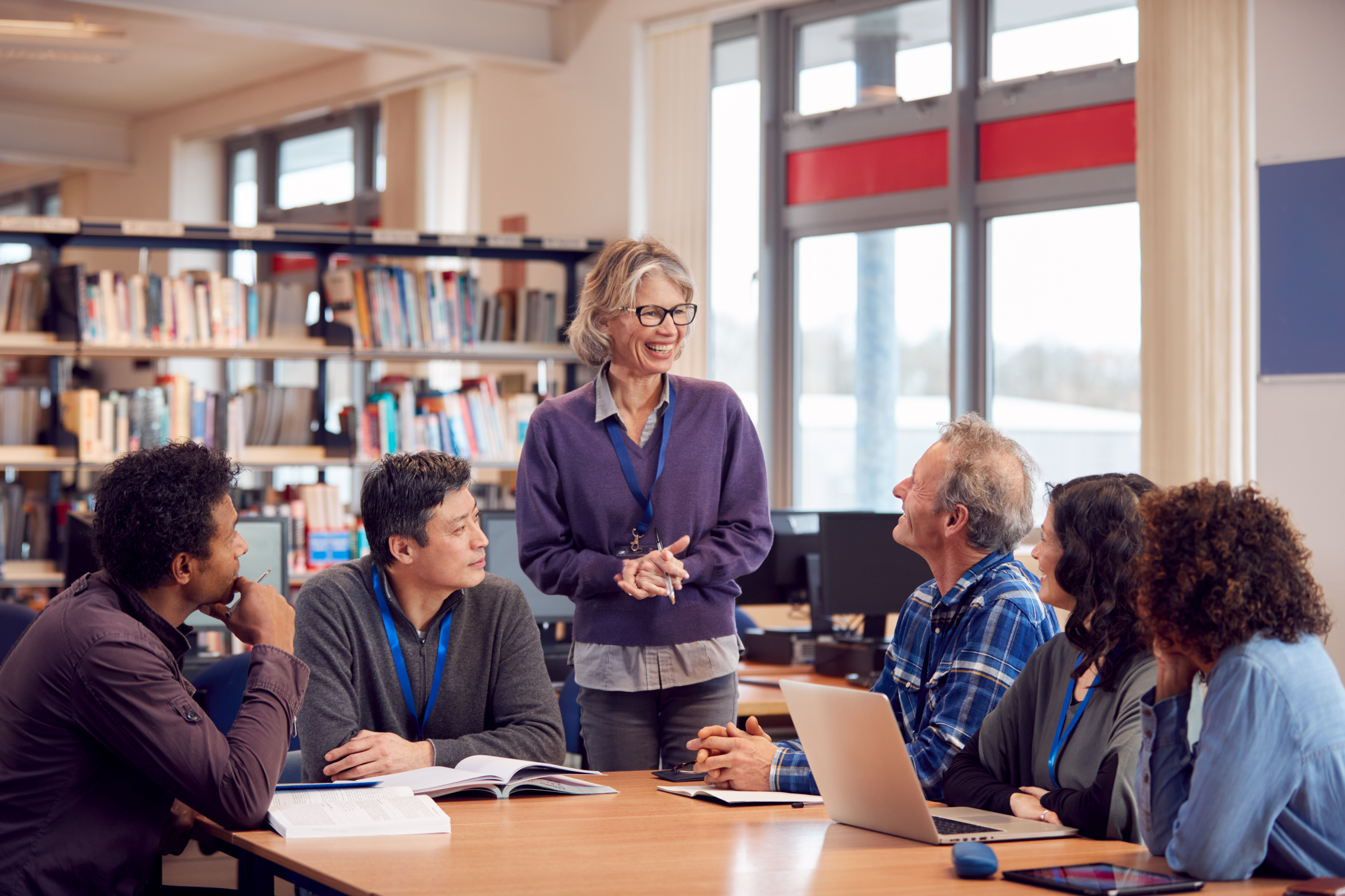 a female teacher standing and leading a discussion with a diverse group of four students or colleagues seated at a table in a library or classroom.