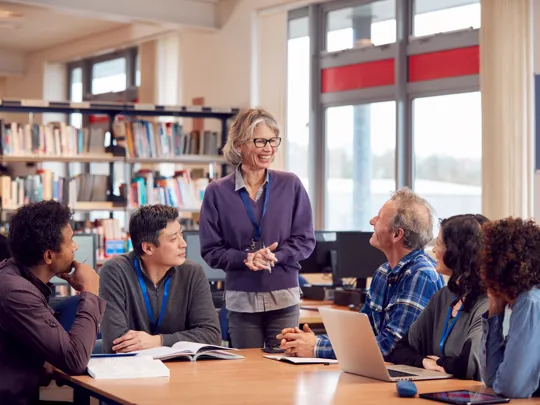 a female teacher standing and leading a discussion with a diverse group of four students or colleagues seated at a table in a library or classroom.
