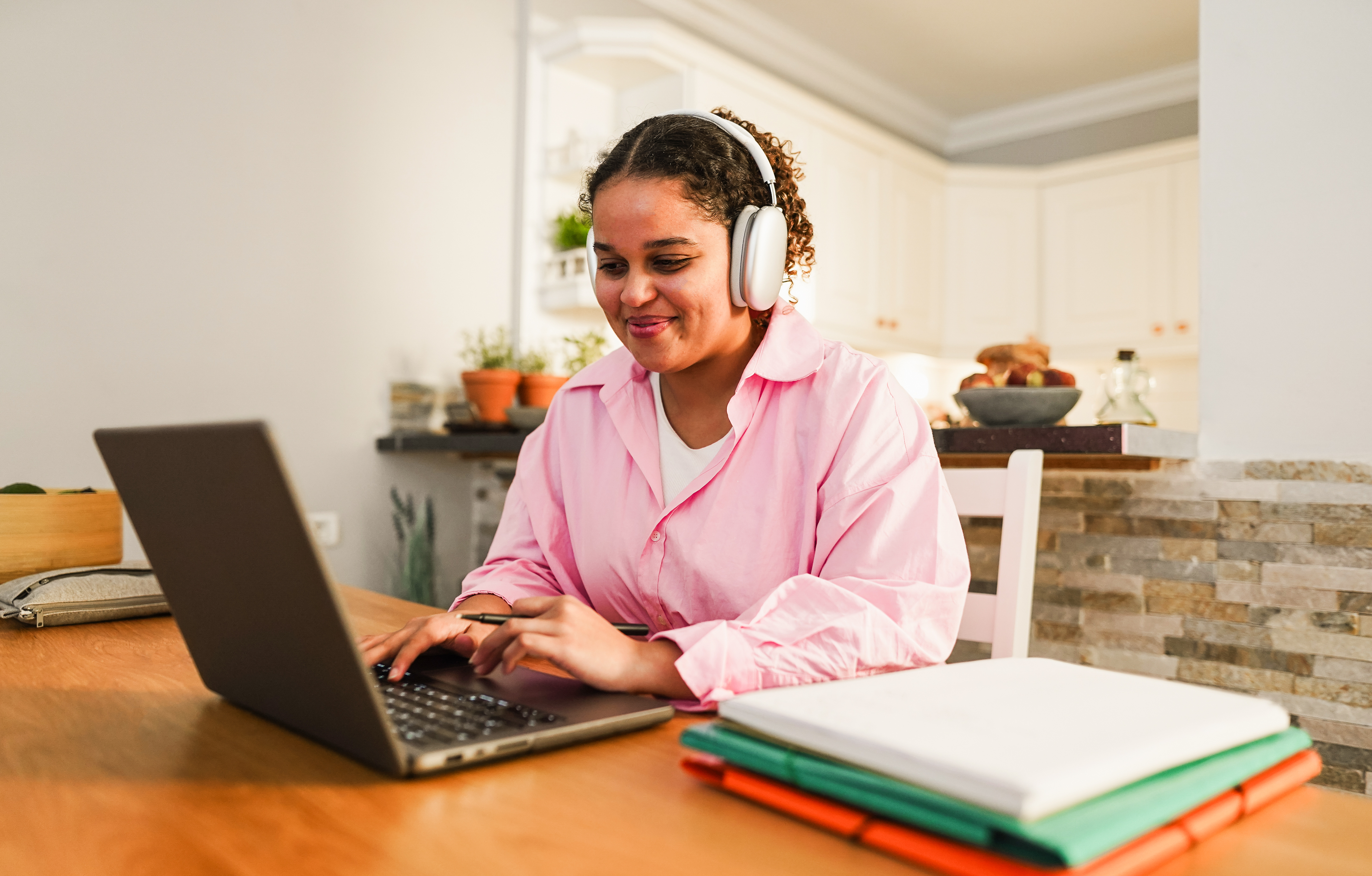 A young female student with curly hair, wearing a pink shirt and white headphones, smiles while typing on a laptop at a kitchen table for online Spanish class.