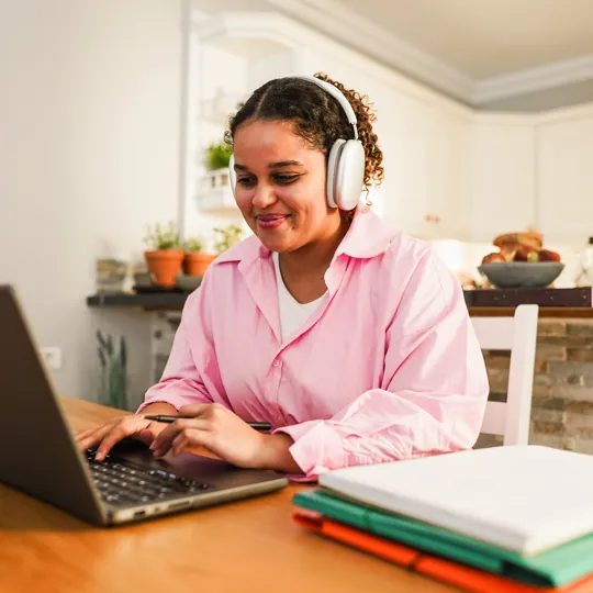 A young female student with curly hair, wearing a pink shirt and white headphones, smiles while typing on a laptop at a kitchen table for online Spanish class.