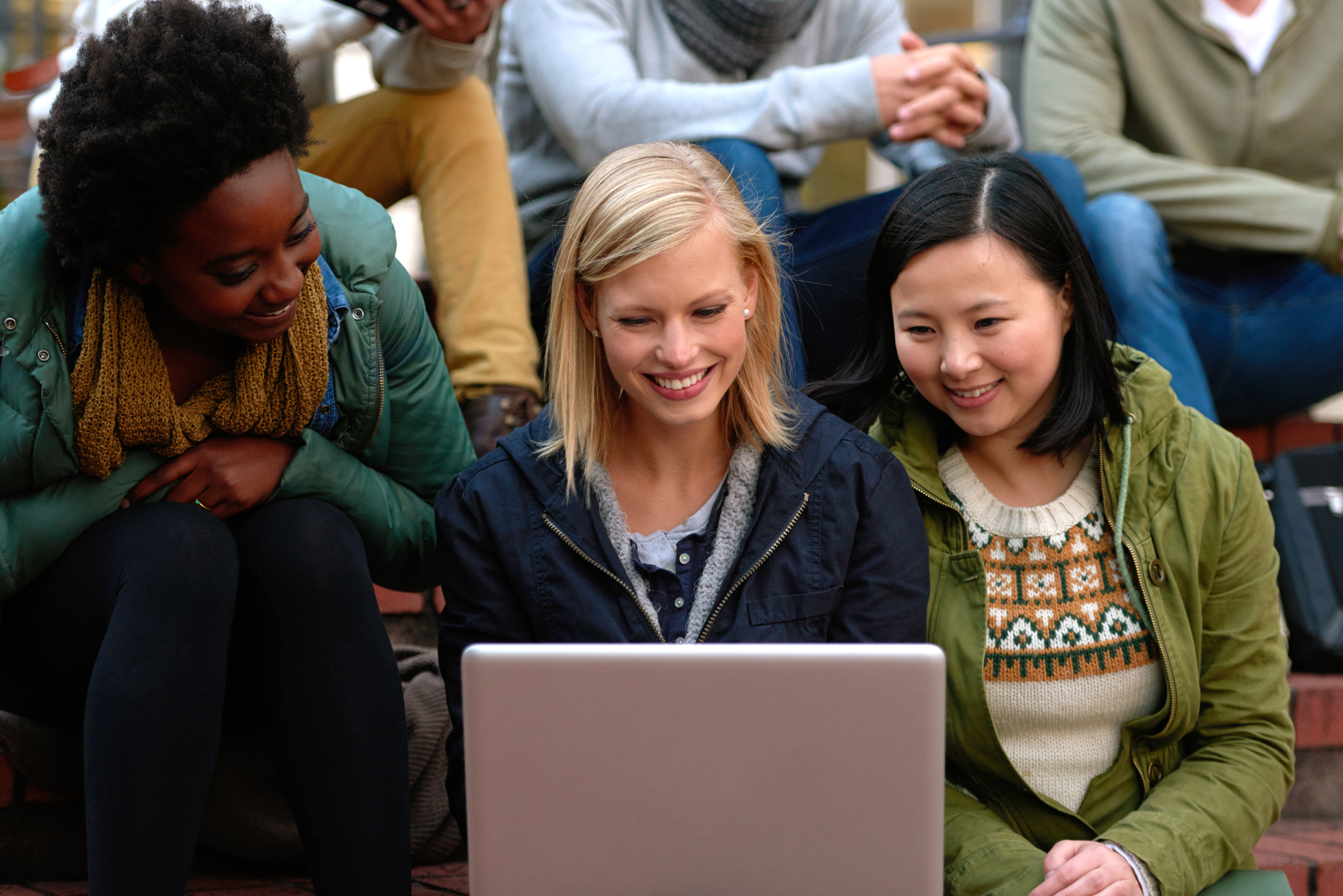 Three smiling young women sitting outside on brick steps looking at a laptop together, with other people sitting behind them.