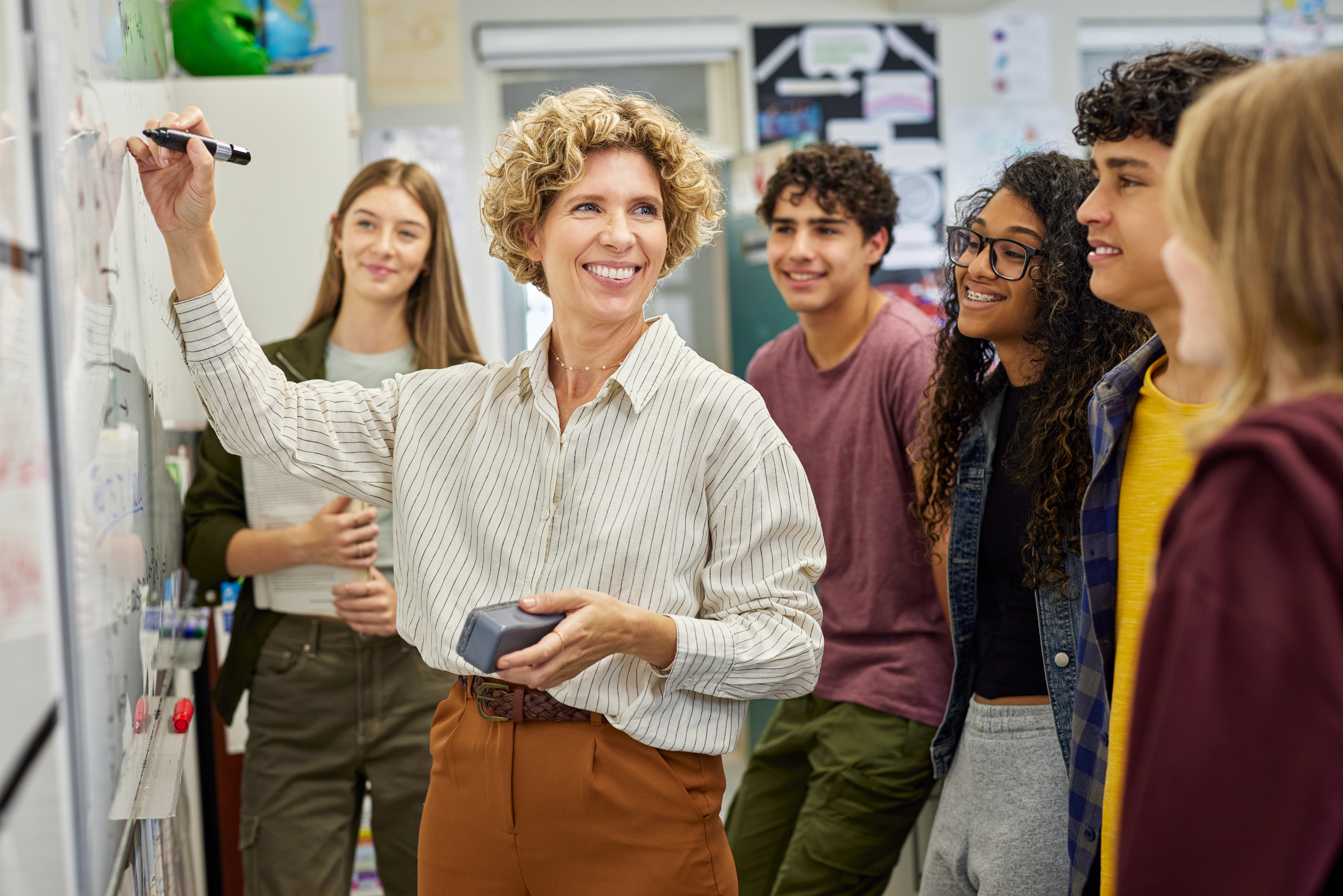 A smiling teacher with curly hair writes on a whiteboard while a diverse group of high school students attentively watches her in a brightly lit classroom.