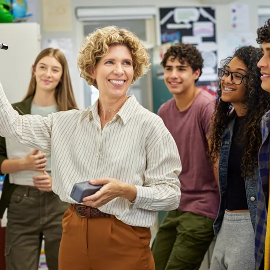 A smiling teacher with curly hair writes on a whiteboard while a diverse group of high school students attentively watches her in a brightly lit classroom.