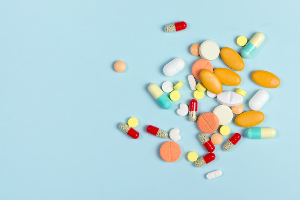 A flat lay of various-sized pills and capsules in different colors, including orange, yellow, red, white, and blue-green, scattered on a solid light blue background.