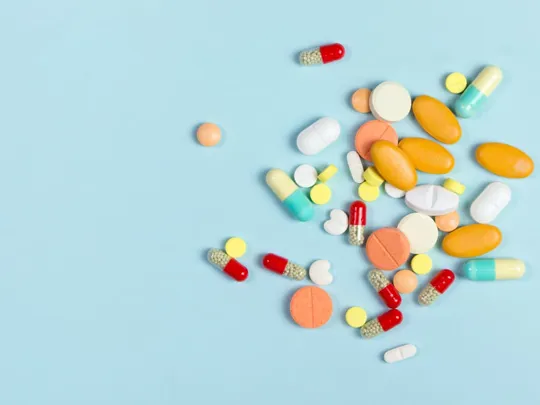 A flat lay of various-sized pills and capsules in different colors, including orange, yellow, red, white, and blue-green, scattered on a solid light blue background.