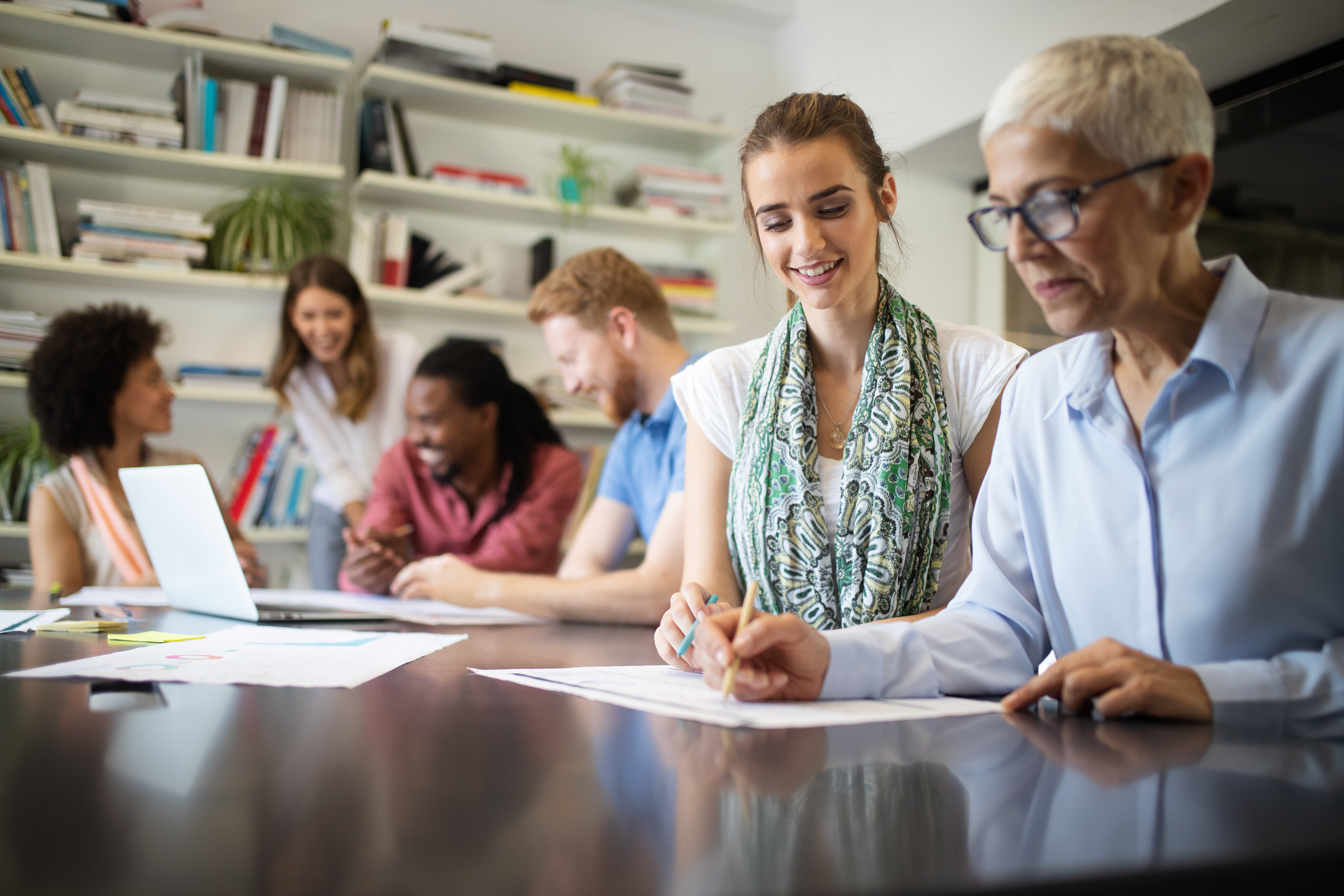 a diverse group of adults working together at a long table in a bright office or library setting with bookshelves in the background.