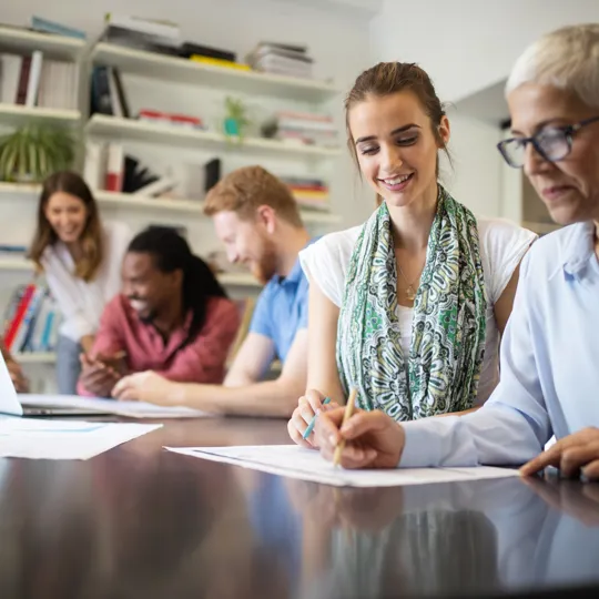 a diverse group of adults working together at a long table in a bright office or library setting with bookshelves in the background.