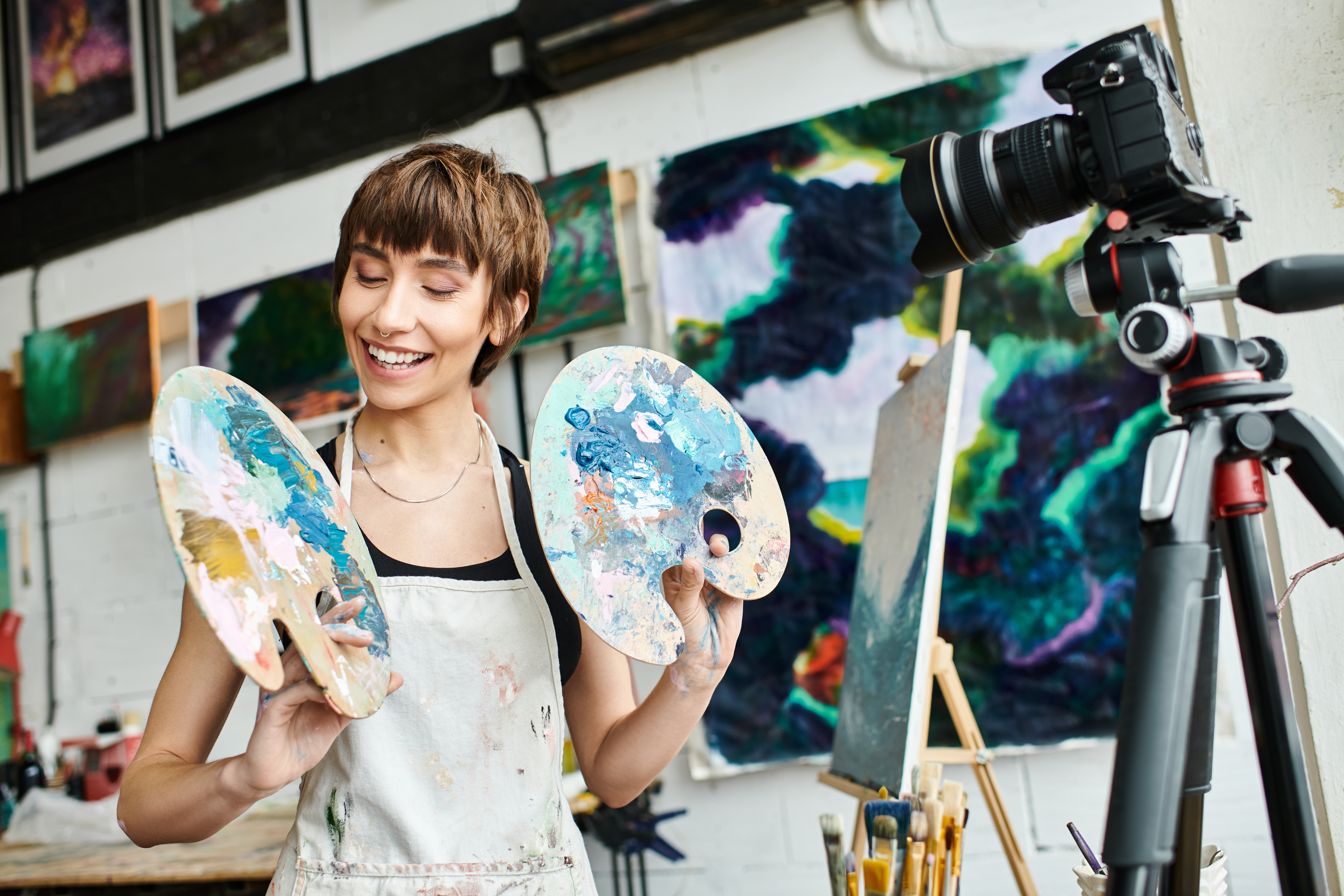 A smiling young woman in an art studio, holding two paint palettes, with a camera on a tripod, easel, and abstract paintings in the background.