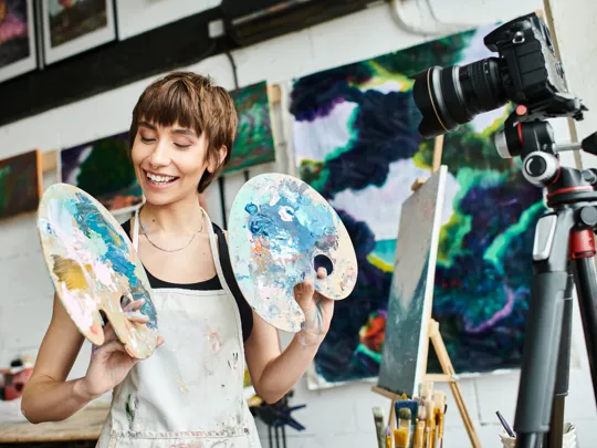 A smiling young woman in an art studio, holding two paint palettes, with a camera on a tripod, easel, and abstract paintings in the background.