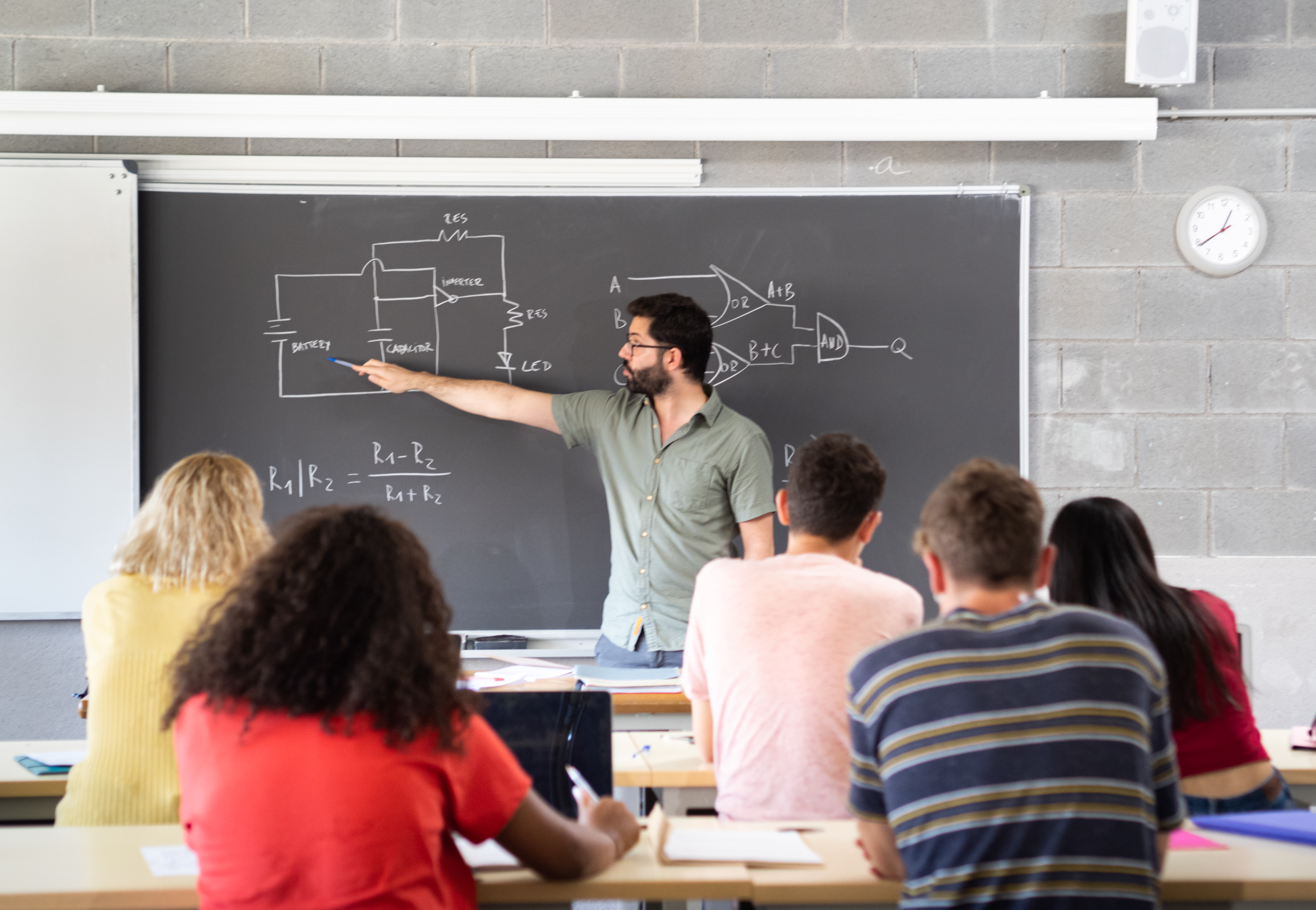a male teacher explaining a circuit diagram on a chalkboard to a class of high school or university students.