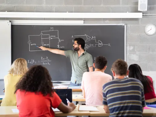 a male teacher explaining a circuit diagram on a chalkboard to a class of high school or university students.