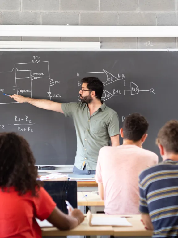 a male teacher explaining a circuit diagram on a chalkboard to a class of high school or university students.