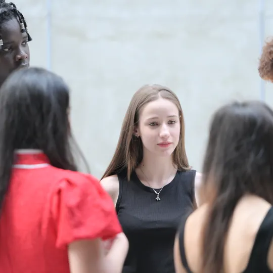 A diverse group of high school students standing in a circle, engaged in a focused discussion or conversation outdoors.