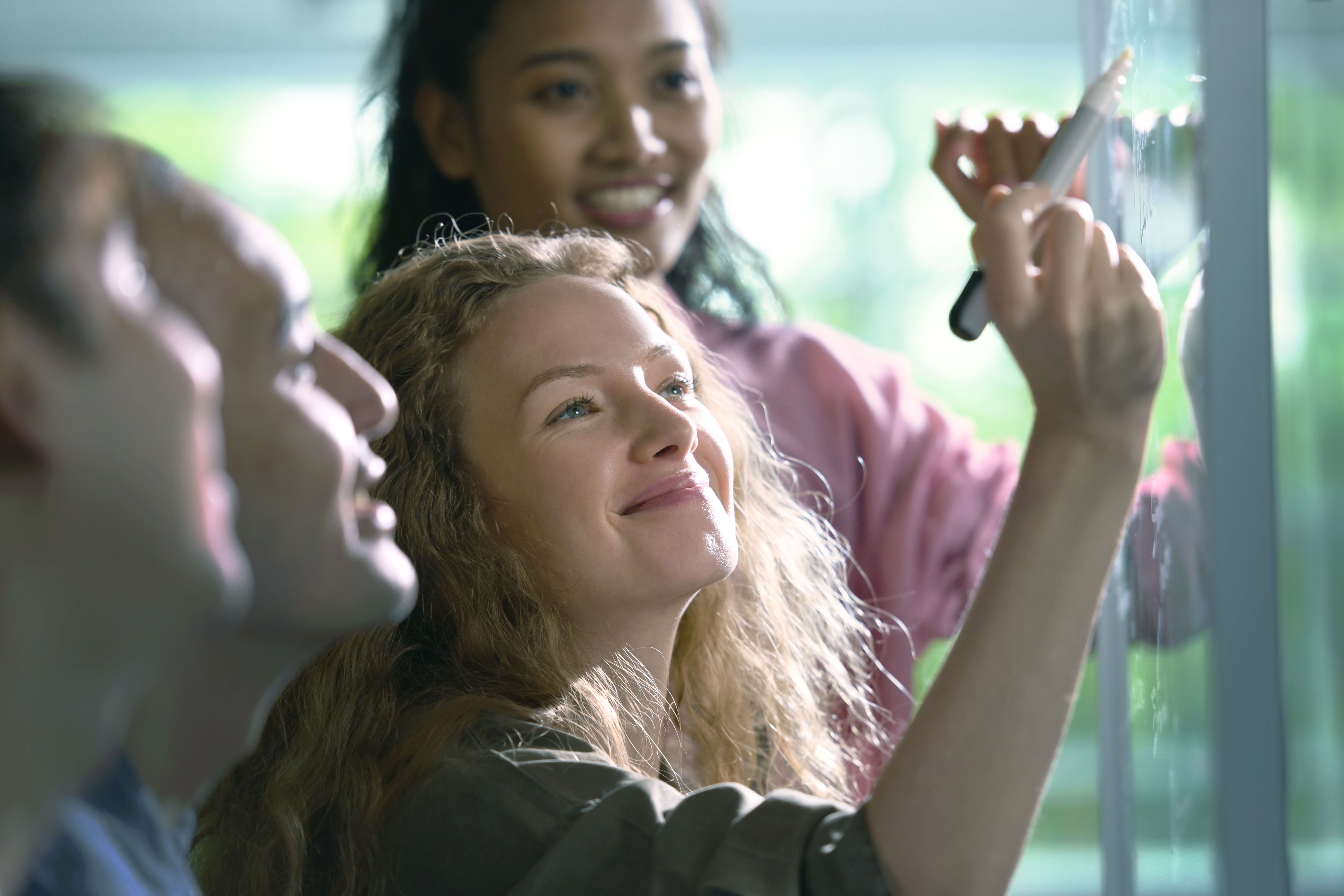 Four smiling colleagues collaborating on an idea; a woman with long blonde hair writes on a clear whiteboard while others look on.