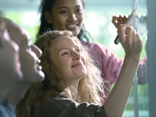 Four smiling colleagues collaborating on an idea; a woman with long blonde hair writes on a clear whiteboard while others look on.