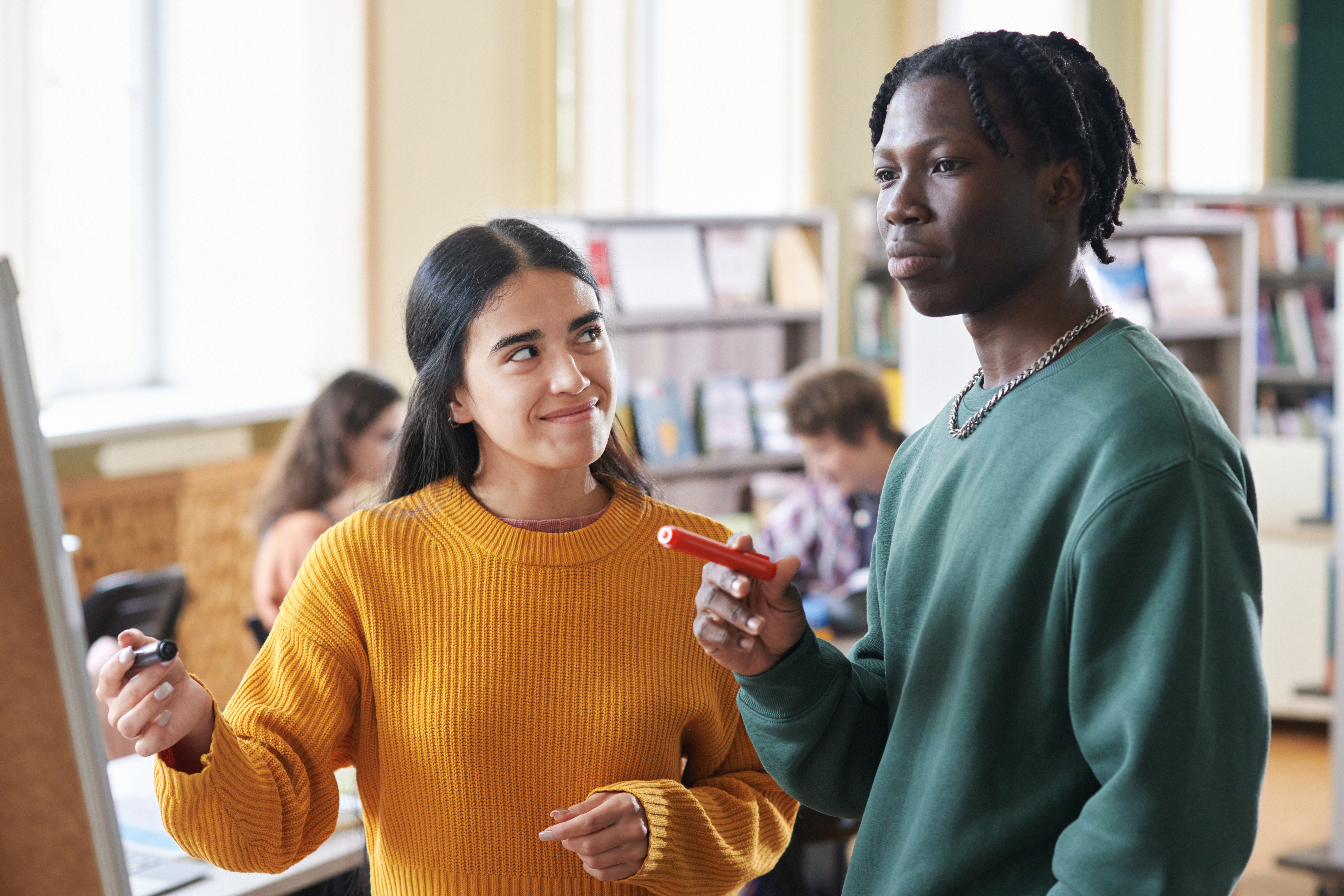 A young man and woman are collaborating on a whiteboard with markers in a bright library or classroom, representing student activities and assessments.