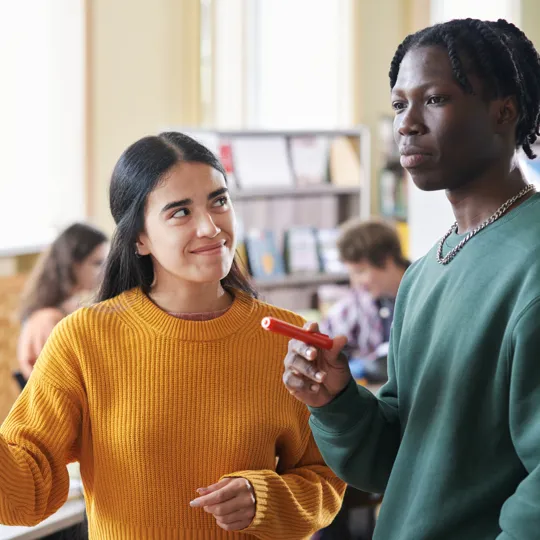 A young man and woman are collaborating on a whiteboard with markers in a bright library or classroom, representing student activities and assessments.